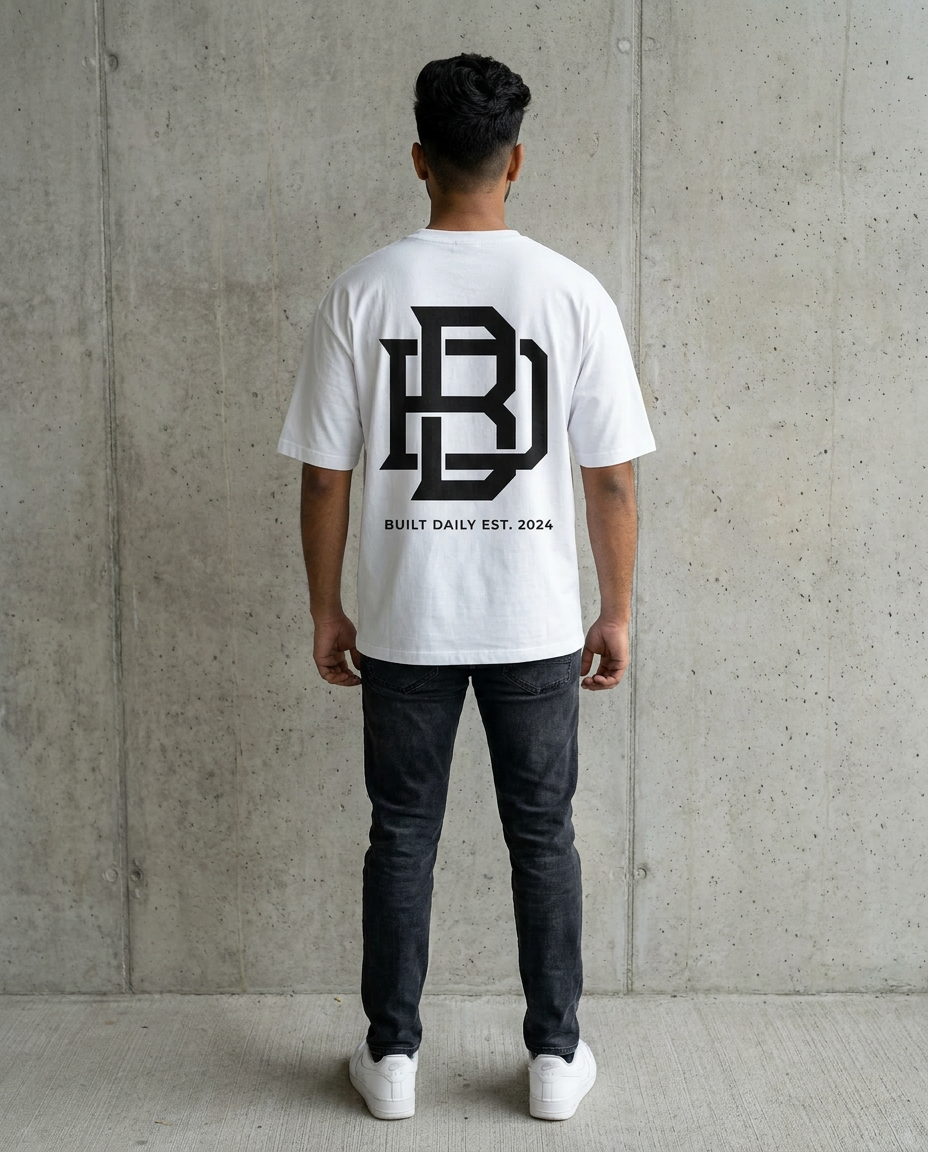 Person wearing a white Oversized t-shirt with a logo and text on the back, standing against a concrete wall.
